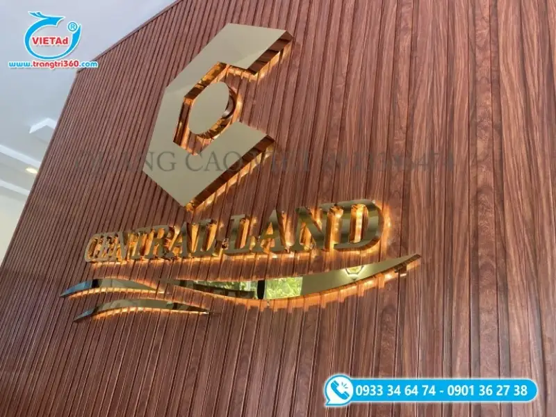 Logo công ty chữ inox vàng hắt sáng trên nền lam gỗ trang trí, tạo phong cách cao cấp cho sảnh công ty